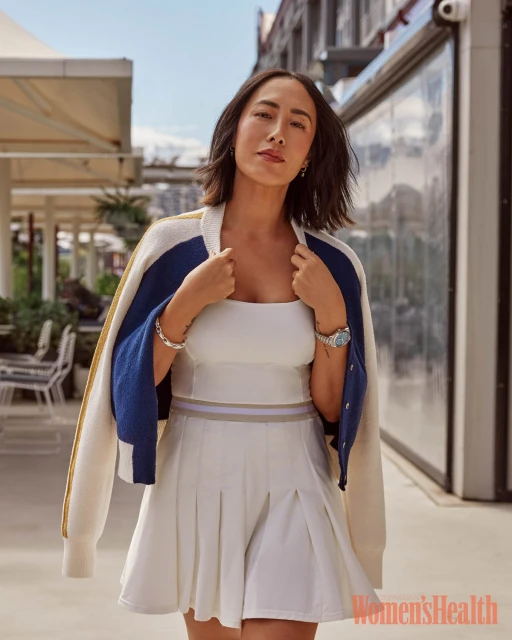 Attractive Asian woman in a tennis outfit poses outdoors with a varsity jacket, in a medium shot with an urban background on a sunny day.